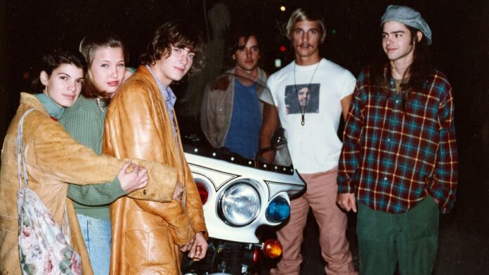 A group of six young adults, embodying a Dazed and Confused vibe, pose casually at night. They're dressed in retro attire, featuring leather and flannel. Standing in front of a classic motorcycle with large headlights, the mood is relaxed and social.