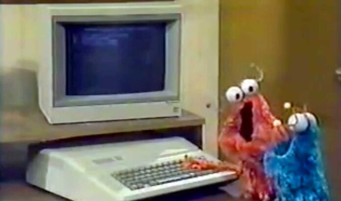 Two fuzzy, wide-eyed puppets, one red and one blue, stand in front of an old computer with a CRT monitor and a keyboard on a desk, appearing curious and animated.