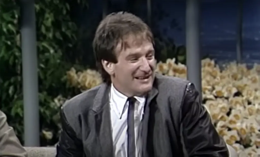 A man with short brown hair wearing a gray jacket, white shirt, and dark tie smiles while sitting on a talk show set decorated with flowers in the background.