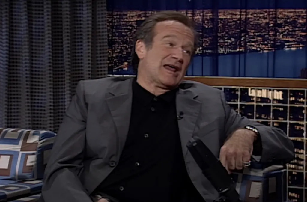 A man in a gray suit and black shirt sits on a patterned couch during a late-night talk show, gesturing expressively and appearing to speak animatedly. The nighttime cityscape is visible in the background.