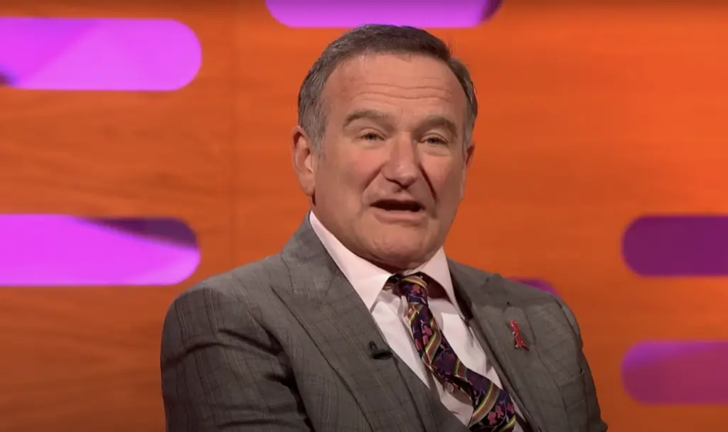 A man in a gray suit and patterned tie sits in front of an orange and purple studio background, speaking or mid-conversation.