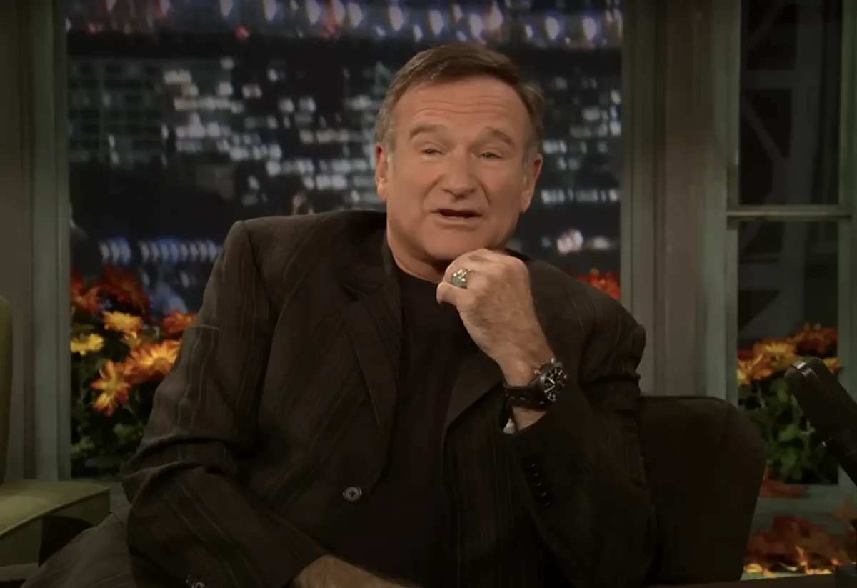 A man in a dark suit and black shirt sits on a talk show set, resting his chin on his hand. The blurred cityscape background features twinkling lights.