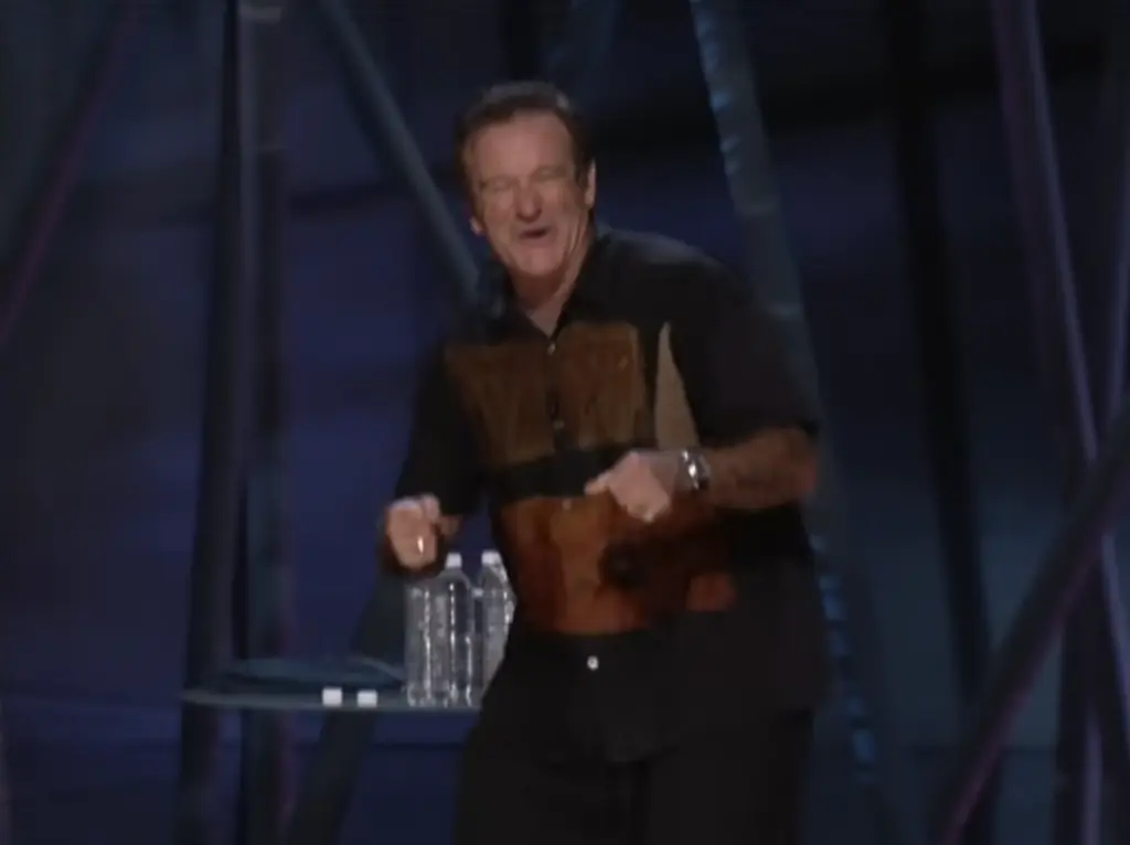 A man wearing a black and brown shirt performs energetically on stage with two water bottles on a small table beside him. The background is dark with structural beams visible.