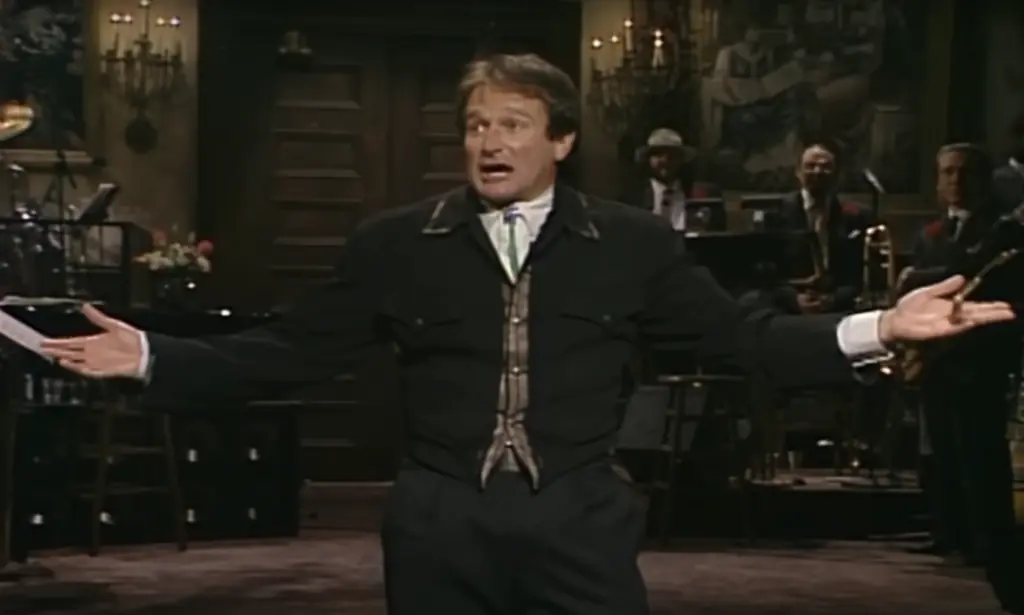 A man in a dark jacket stands on a stage with his arms spread wide, speaking animatedly. Musicians and instruments are visible in the dimly lit background.