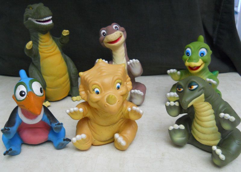 Toy dinosaurs sit cheerfully on a surface with a dark background, reminiscent of a playful Pizza Hut party. They appear colorful and cartoon-like with exaggerated features. Each has a unique design and expression, showcasing a variety of vibrant colors, bringing dino joy to the table.