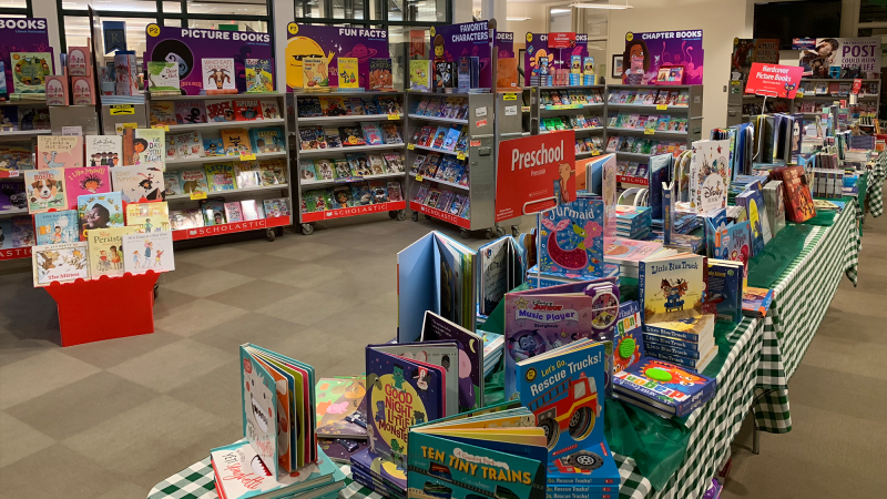 A colorful book fair display with tables and shelves filled with childrens books. Sections are labeled, including Picture Books, Fun Facts, and Preschool. The tables have green checkered tablecloths.