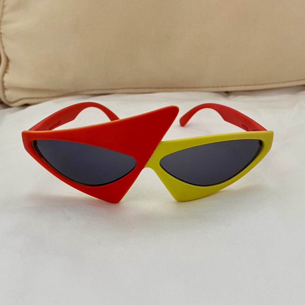 A unique pair of sunglasses with an asymmetrical design, reminiscent of Pizza Hut's bold flair. The left lens is framed in red with a large triangular shape, while the right is in yellow with a smaller triangle. The arms are strikingly red, adding to their standout charm.