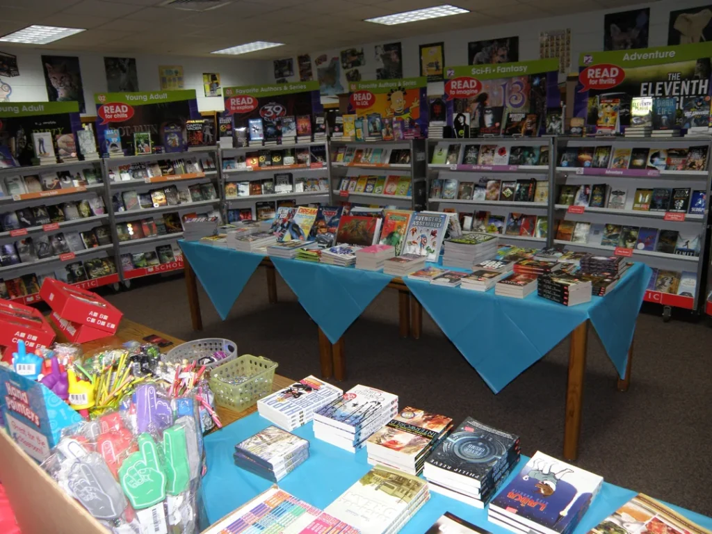 A vibrant book fair with tables covered in blue tablecloths displaying a variety of books. Shelves lined with books in sections such as young adult, fantasy, and adventure. A basket of colorful stationery is in the foreground.