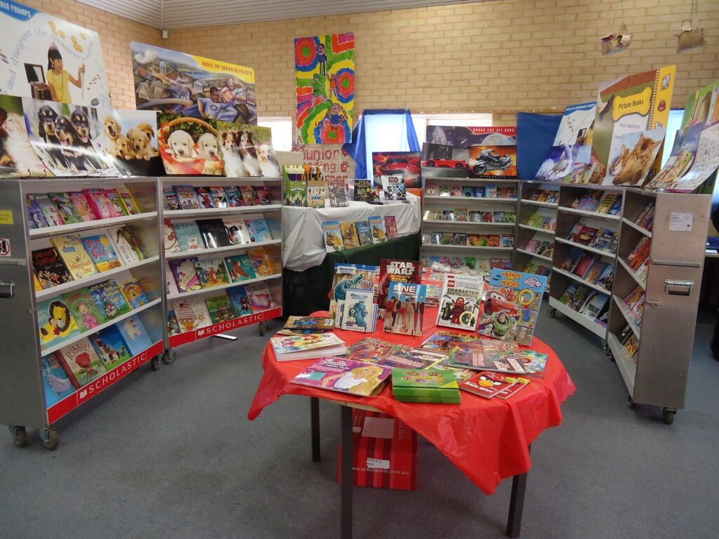 A colorful book fair display features shelves filled with childrens books. In the center, a table covered with a red cloth holds various books, including titles like Star Wars. Stands and posters surround the area, creating an inviting atmosphere.