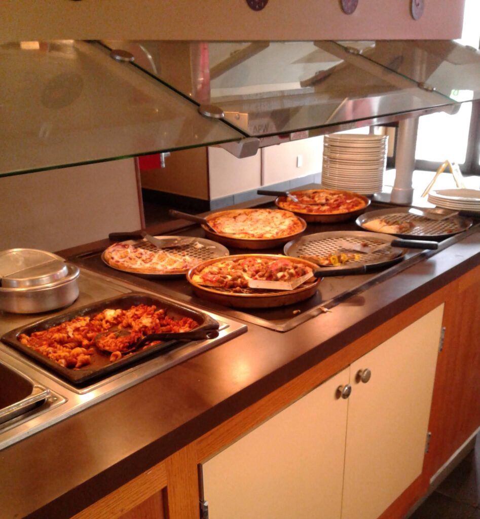 A buffet line resembling a Pizza Hut feast showcases multiple pizzas with various toppings, including pepperoni and vegetables, alongside trays of pasta dishes. Plates are neatly stacked on the right side under a glass sneeze guard.