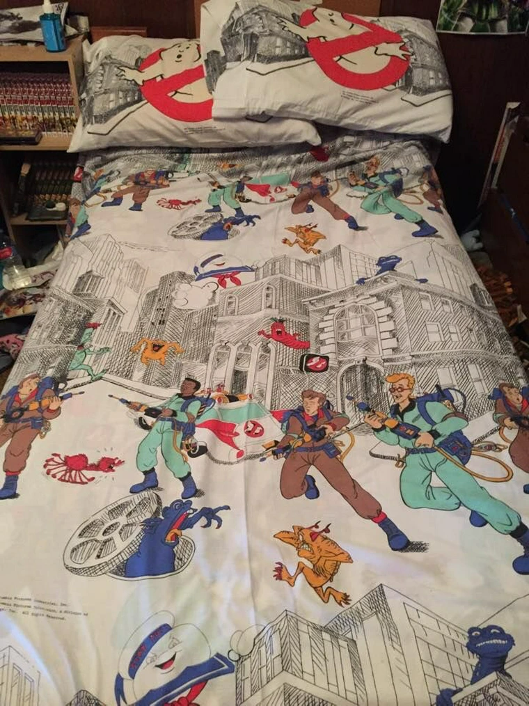 A bed with Ghostbusters-themed bedding features characters in action poses against a cityscape, surrounded by ghosts and paranormal elements. Illustrated pillowcases and sheets amplify the theme, resembling a treasure trove of Ghostbusters collectibles in a cluttered room.