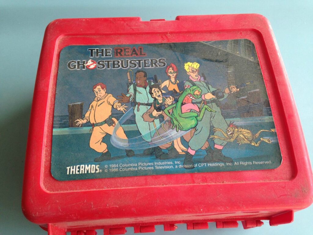 A red lunchbox featuring The Real Ghostbusters cartoon characters in action, showcasing their ghost-catching gear and a friendly green ghost. This Ghostbusters Collectible includes the show's logo and copyright details. The lunchbox is slightly worn, adding vintage charm.