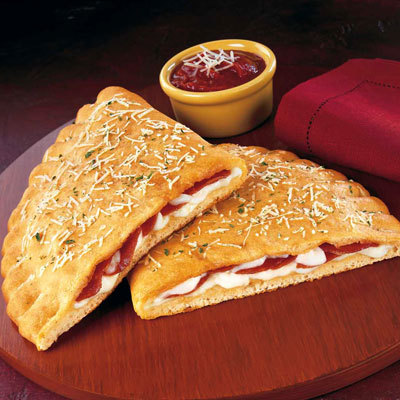 A sliced calzone filled with pepperoni and melted cheese sits on a wooden board, reminiscent of a Pizza Hut creation. The golden-brown crust is sprinkled with herbs and grated cheese. A small bowl of marinara sauce and a folded red napkin are next to it.