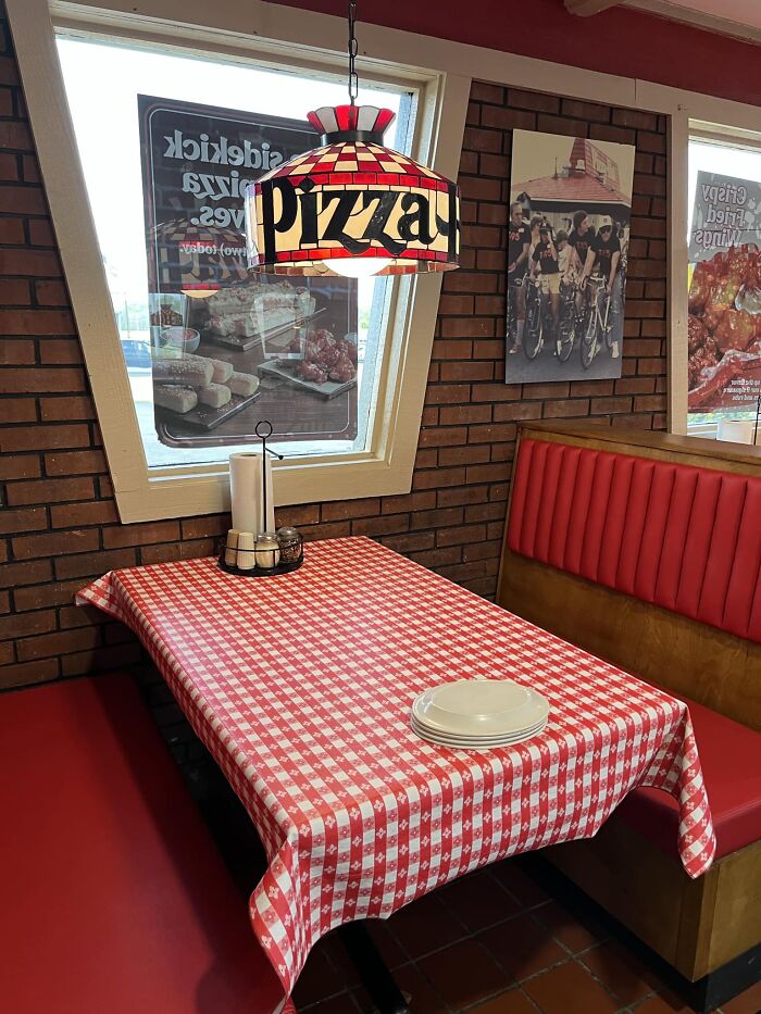 A cozy Pizza Hut booth featuring red leather seating and a table with a red and white checkered tablecloth. A vintage-style lamp with "pizza" written on it hangs above. Walls are decorated with posters and brickwork.