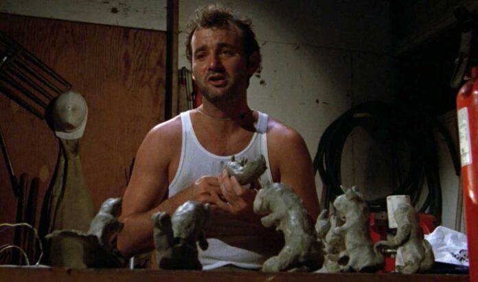 Bill Murray in Caddyshack in a white tank top sits at a table in a dimly lit room, surrounded by clay sculptures. A fire extinguisher and a straw hat are visible in the background. Channeling Bill Murray quotes from Caddyshack, the person is deeply engaged with the clay figures.