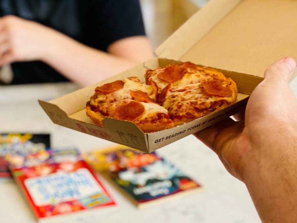 A hand holds an open Pizza Hut box with a personal-sized pepperoni pizza. In the background, another person sits at a table, three colorful comic books scattered in front of them.