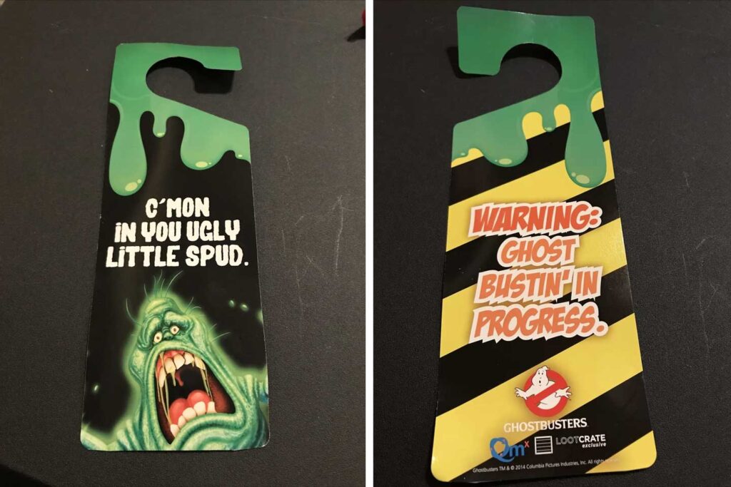 Two door hangers: one flaunts green slime and a ghost with "CMON IN YOU UGLY LITTLE SPUD." The other showcases black/yellow stripes reading "WARNING: GHOST BUSTIN IN PROGRESS," accompanied by the Ghostbusters logo. Perfect for fans of Ghostbusters Collectibles.