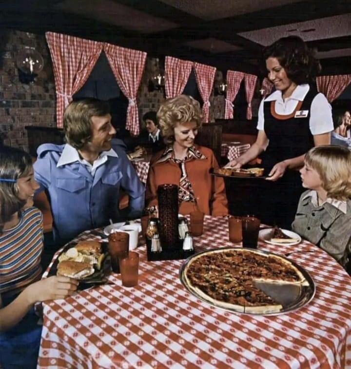 A family of four sits at a table with a checkered tablecloth, savoring delicious Pizza Hut pizza and burgers. A waitress serves them with a smile. The restaurant ambiance is cozy, featuring checkered curtains and dim lighting, perfect for a delightful dining experience.