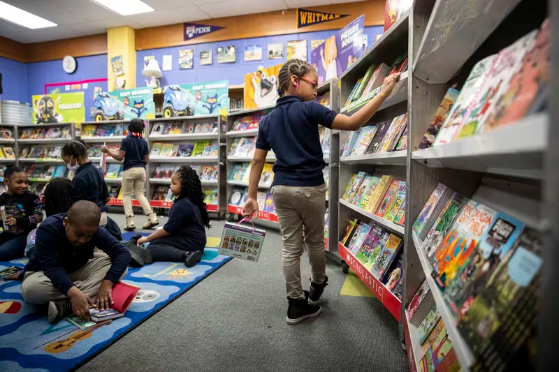 Children browse and read books in a colorful library. Some sit on a patterned carpet, while others stand by shelves filled with vibrant book covers. The atmosphere is lively and engaging, fostering a love for reading.