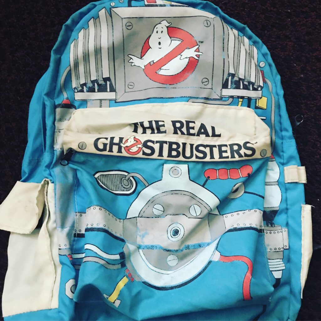 A blue backpack featuring The Real Ghostbusters logo and design, including an illustration of the Ghostbusters emblem and machinery graphics. This piece of Ghostbusters collectibles shows signs of wear.