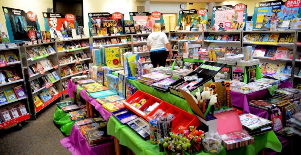 A colorful book fair display with tables covered in books, stationery, and school supplies. Shelves in the background hold more books and posters. A person browses through the items. Bright banners and signs create a lively atmosphere.