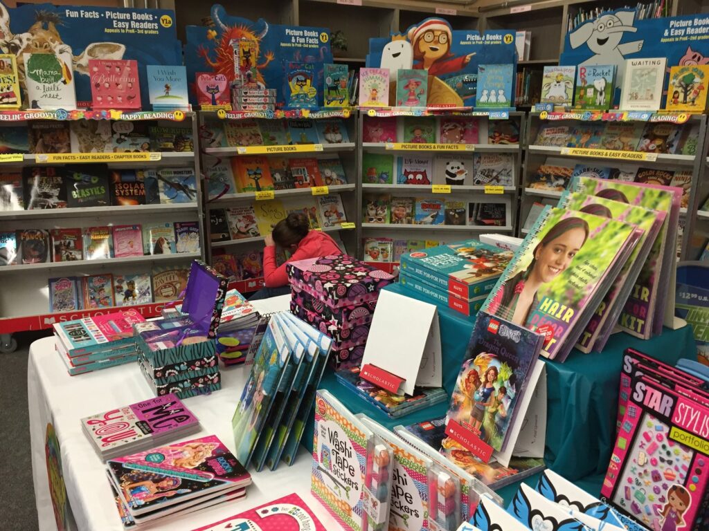 A child in a red jacket browses books in a colorful book fair. The shelves are stocked with childrens books, including storybooks and educational titles. A table in the foreground displays more books alongside school supplies.