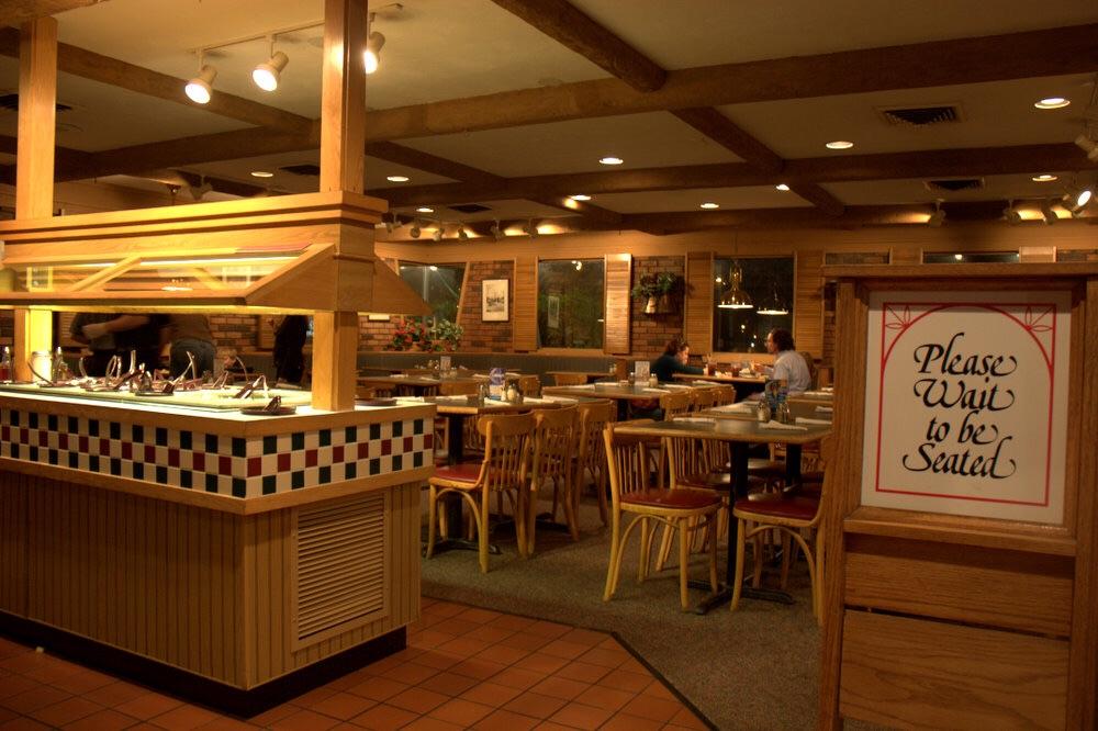 The interior of a warmly lit restaurant, reminiscent of Pizza Hut's inviting vibe, features wooden furniture. A buffet setup is on the left, and a sign near the entrance reads Please Wait to be Seated. Several tables are visible, with a few customers dining.