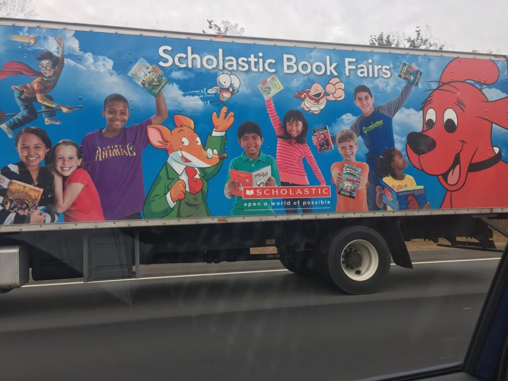 A truck displays a colorful Scholastic Book Fairs advertisement with children and illustrated characters holding books. The background is bright blue, featuring the slogan open a world of possible and the Scholastic logo.