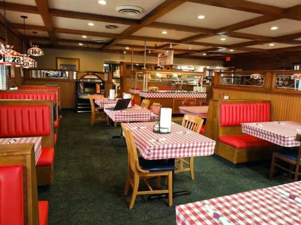 A cozy diner reminiscent of a classic Pizza Hut, featuring red booths and wooden chairs around tables with red and white checkered tablecloths. Wooden beams line the ceiling, and pendant lights hang above the seating areas, creating an inviting atmosphere.