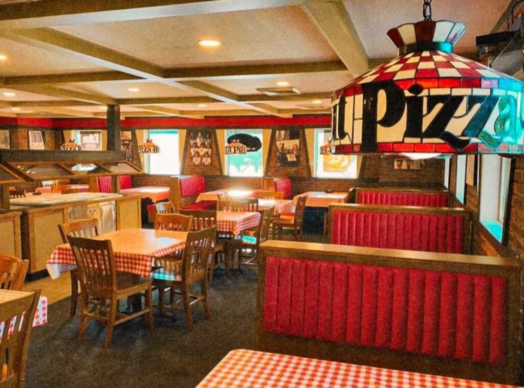 A cozy Pizza Hut restaurant interior with red and white checkered tablecloths, wooden chairs, and red booth seating. Stained glass pendant lights hang above, and framed pictures decorate the walls, creating a warm, inviting atmosphere.