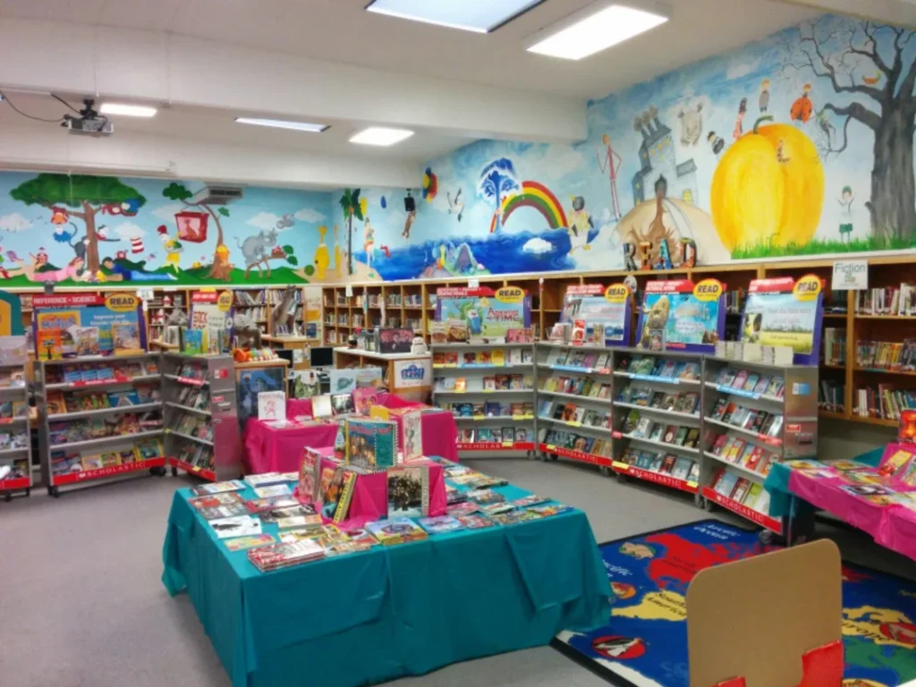 A colorful bookstore with shelves of childrens books. The walls feature vibrant murals of trees, animals, and a rainbow. Tables with books are set up in the center, and the floor has a brightly colored alphabet rug.