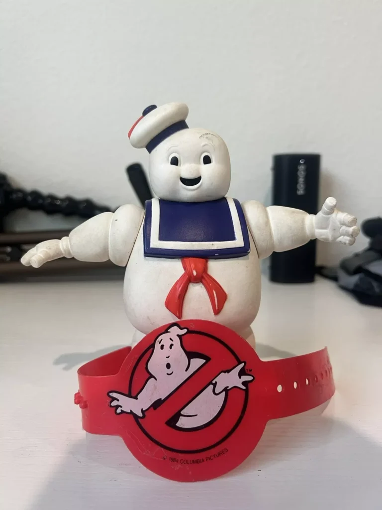 A small plastic figure of the Stay Puft Marshmallow Man stands on a white surface, smiling with open arms. In front of it is a red Ghostbusters-themed watch featuring the iconic no-ghosts logo, making this scene a charming addition to any Ghostbusters collectibles display.