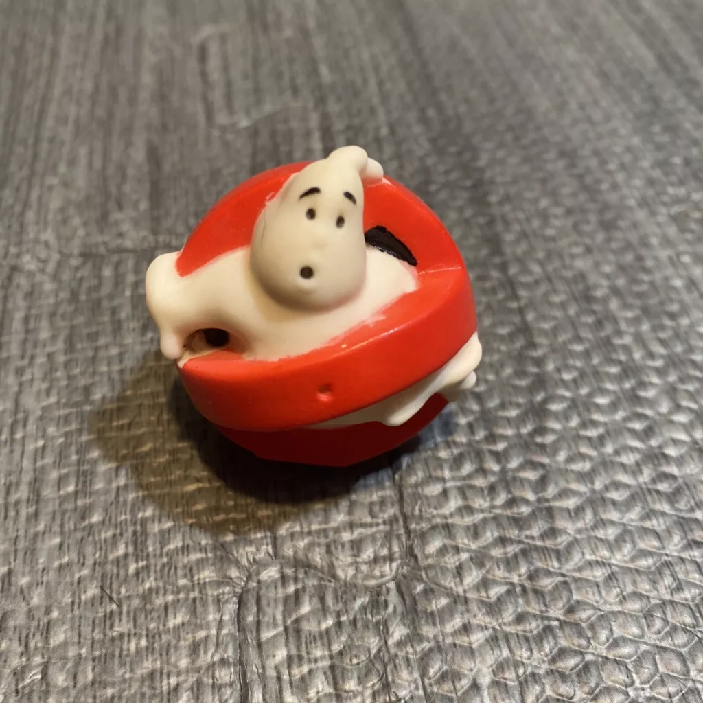 A small toy or model of a white ghost emerging from a red circle with a horizontal bar, resembling the iconic Ghostbusters logo, rests on a textured gray surface—an ideal addition for fans and collectors of Ghostbusters collectibles.