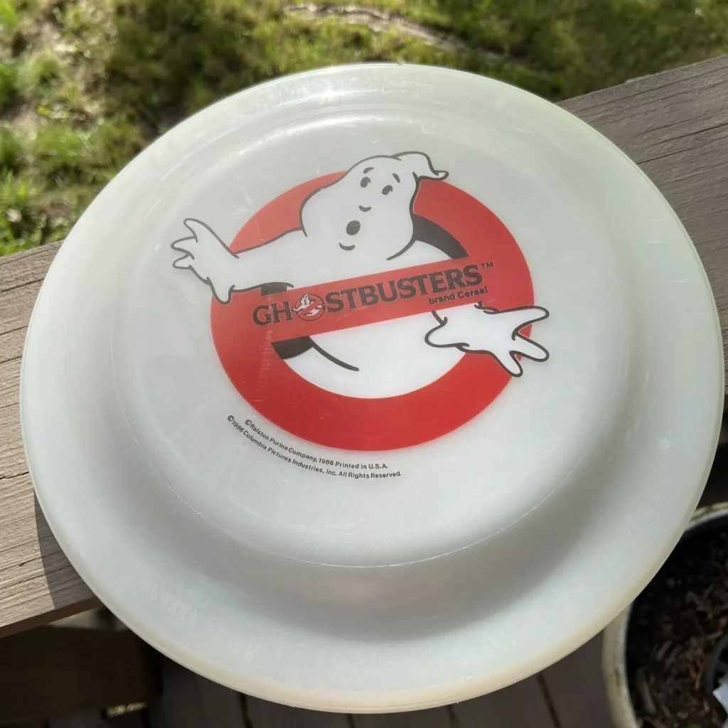 A white frisbee featuring the iconic Ghostbusters logo—a cartoon ghost behind a red prohibition symbol—rests on a wooden surface with grass peeking through in the background. This piece is perfect for any Ghostbusters collectibles enthusiast.