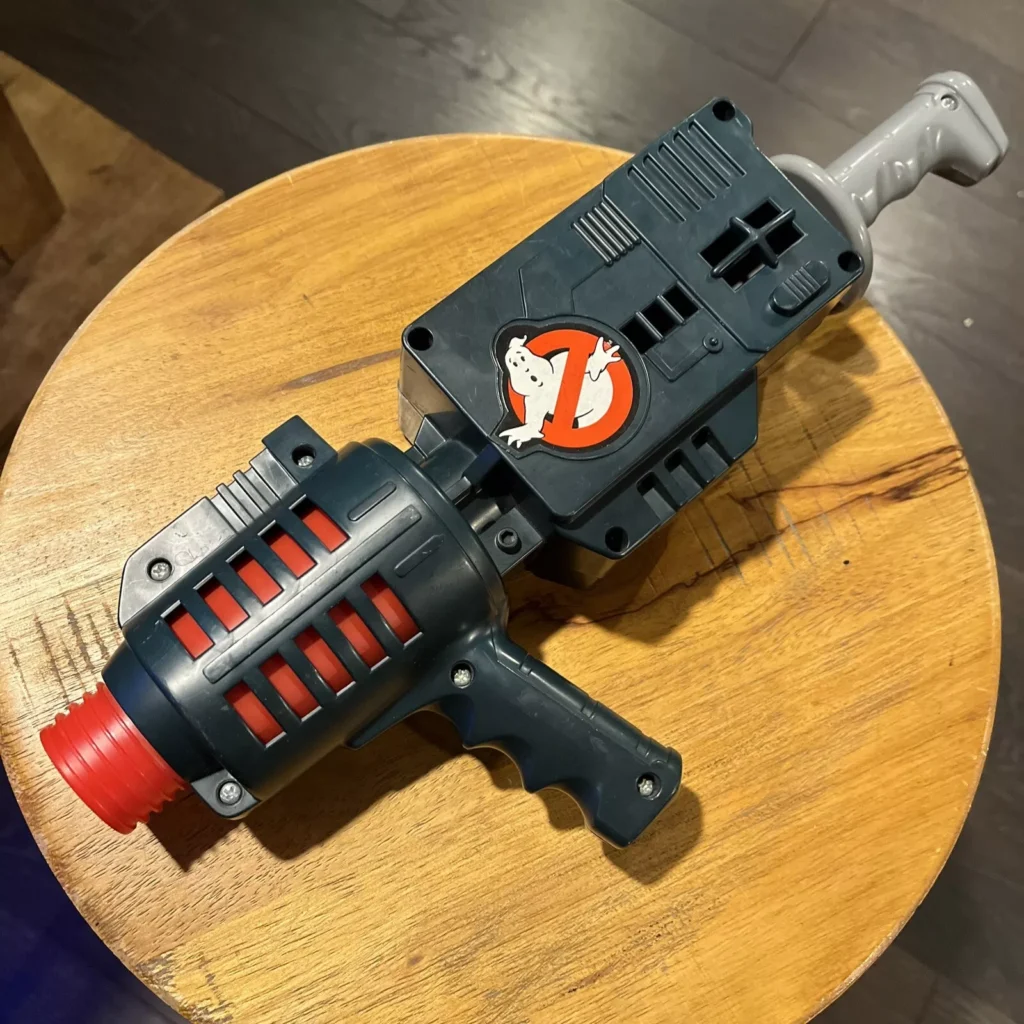 A toy proton pack blaster from Ghostbusters Collectibles rests on a wooden surface. It features a black body with red and gray accents, including a handle and the iconic Ghostbusters logo.