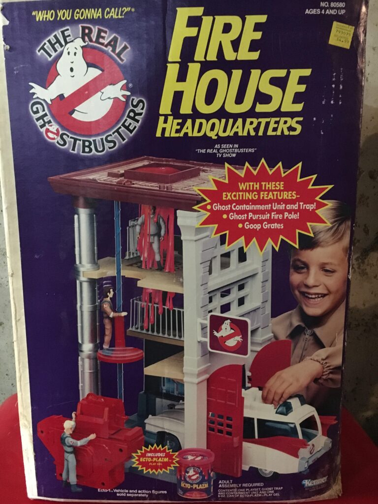 Front cover of The Real Ghostbusters Fire House Headquarters toy box. The image highlights a child playing with this multi-level playset, complete with a containment unit, trap, and Ecto-1 vehicle. It's a must-have for fans of Ghostbusters Collectibles in bright red and blue packaging with promotional text.