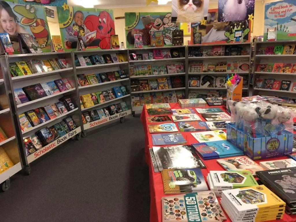 A bustling book fair featuring shelves of colorful childrens books and toys. Books are displayed on tables and shelves, with vibrant posters and decorations overhead. The setting has a lively and inviting atmosphere.