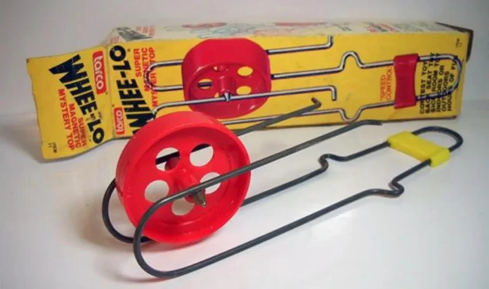 A vintage toy, the Whee-lo, is displayed. This nostalgic treasure has a red plastic wheel on a metal track designed like a handle. In the background, the box showcases vibrant retro packaging and illustrations that capture its playful charm.