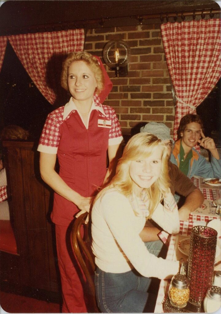 A waitress in a red and white uniform stands by a table with gingham tablecloths, embodying the classic charm of Pizza Hut. Three people are seated, smiling at the camera in this cozy restaurant setting, with pizza condiments scattered invitingly across the table.