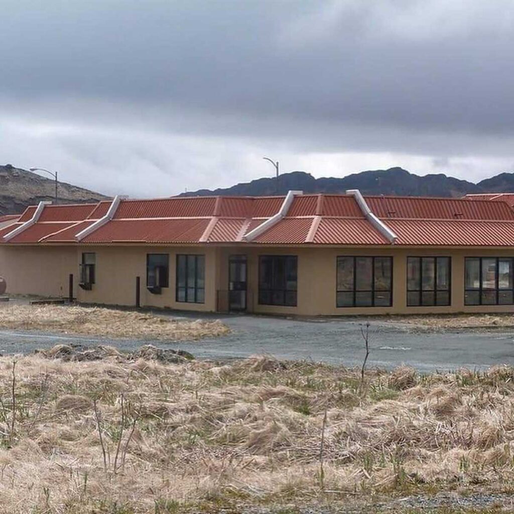 A building with a red-tiled roof, beige walls, and large windows surrounded by a grassy field. Hills are visible in the background under a cloudy sky.