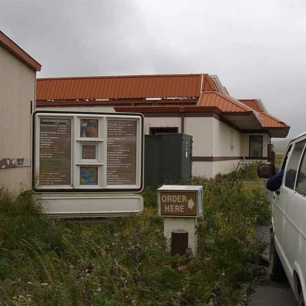 An abandoned McDonald's drive-thru with overgrown grass leading to a rusty building with an orange roof. A weathered sign says ORDER HERE near a white van parked on the right. The menu board is old and faded.