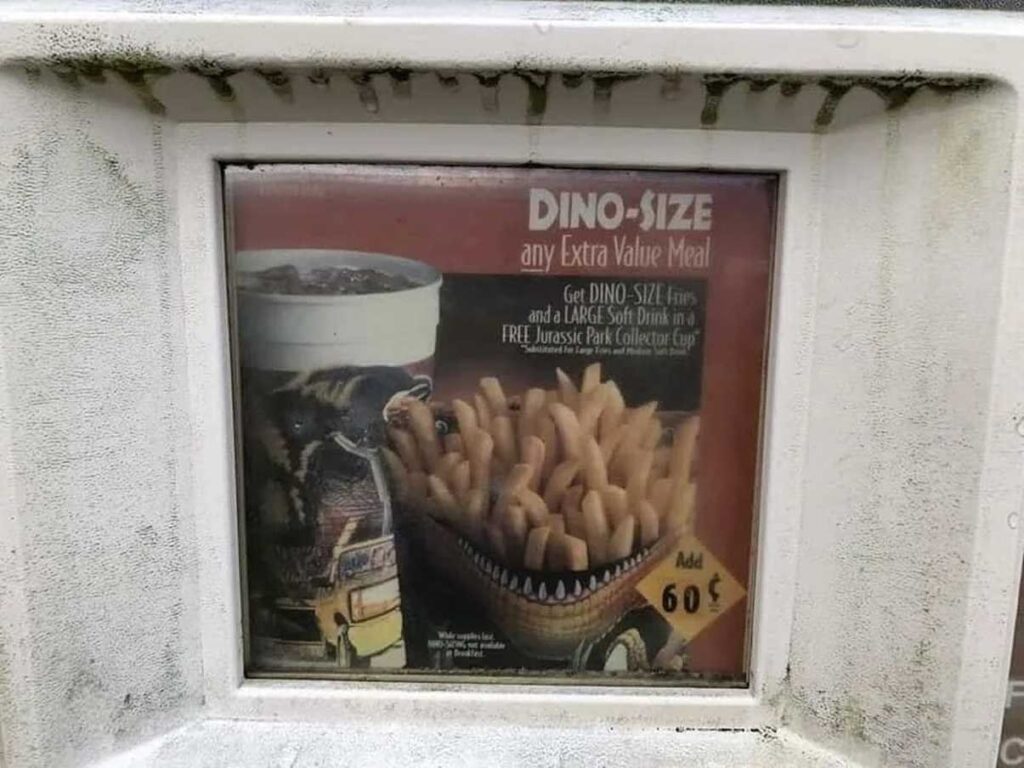 A faded, weathered advertisement for a fast-food promotion offers an option to upgrade to Dino-Size an extra value meal, featuring a large cup and fries for an additional cost of 60 cents. The ad includes a dinosaur graphic.