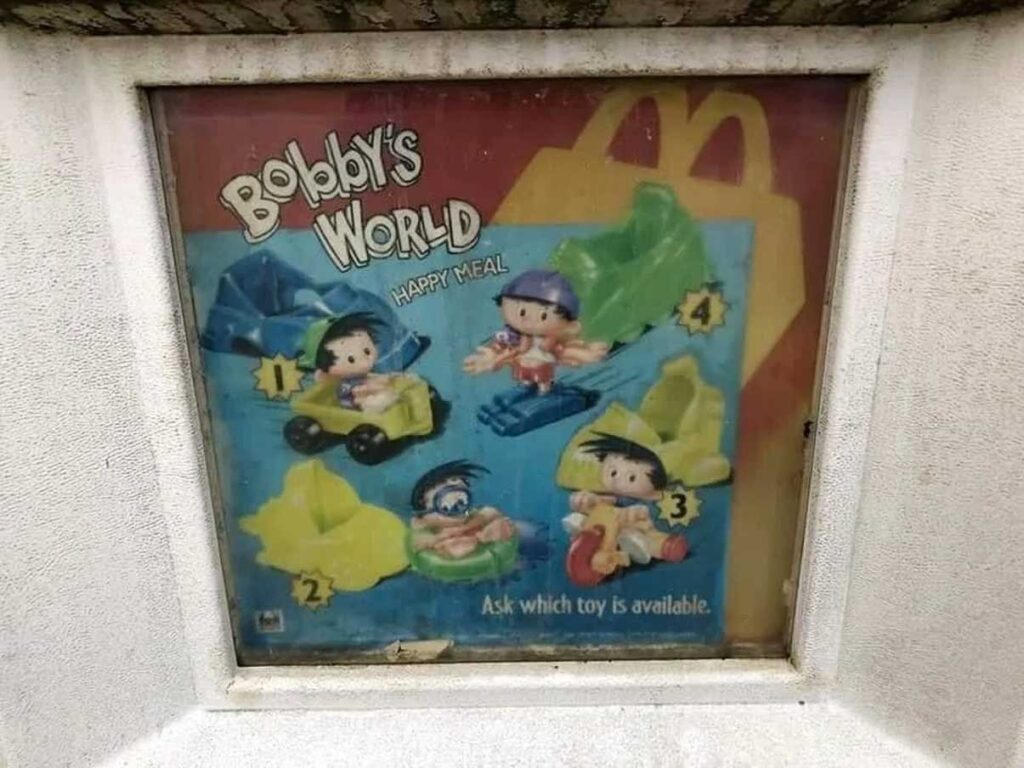 An old McDonalds display for Bobbys World Happy Meal toys shows four toy figures in different vehicles, labeled 1 to 4. The toys are cartoon characters sitting in various colorful cars and planes.