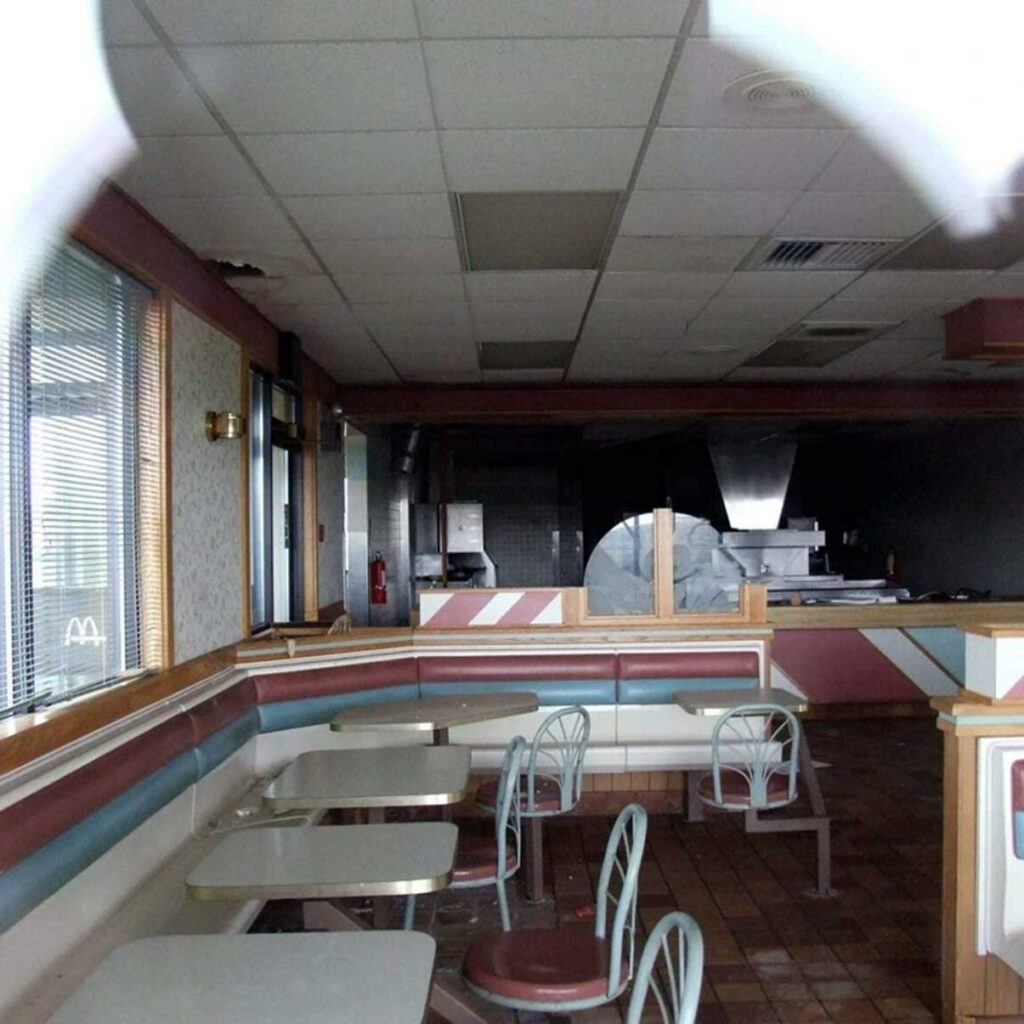 Empty fast-food restaurant interior with pastel-colored booths and tables. The chairs are neatly arranged, and the counter area is visible in the background. The lighting is dim, and the windows show closed blinds.