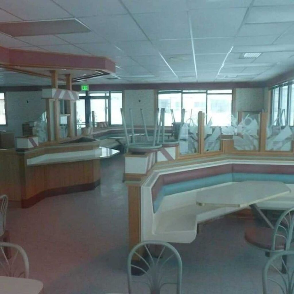 An empty fast-food restaurant interior with pastel-colored booths and tables. Chairs are stacked upside-down on some tables, and natural light comes in through the large windows. The decor features geometric patterns and a retro style.