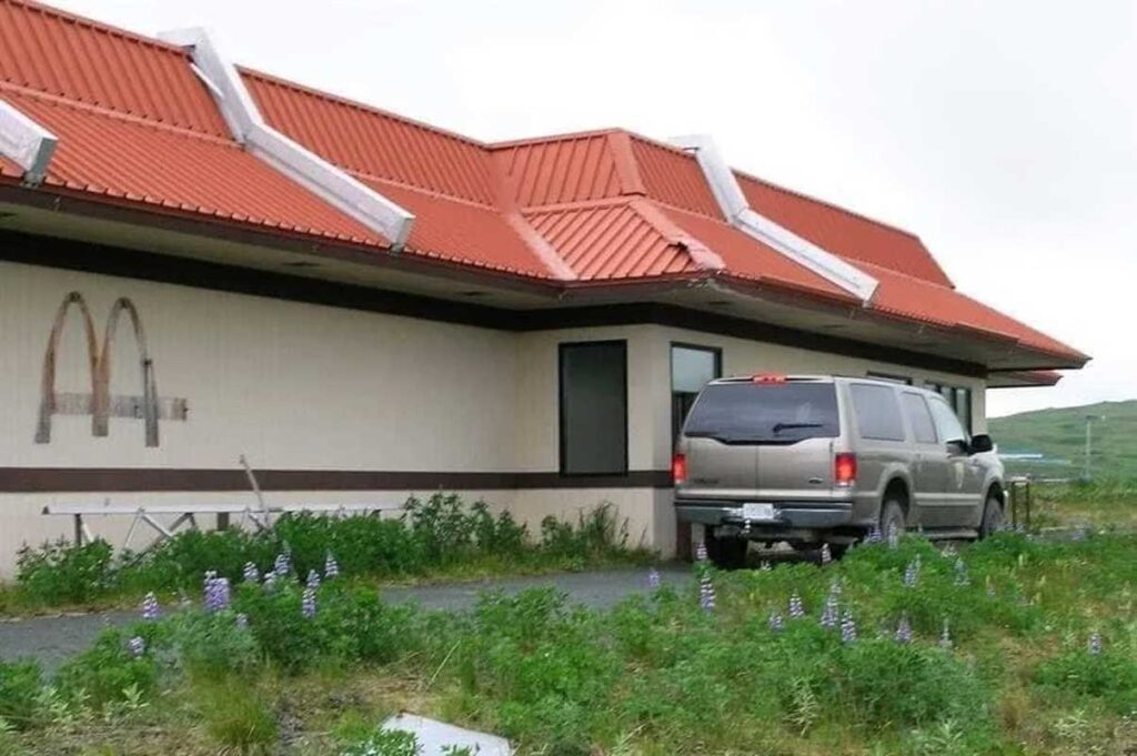 An abandoned fast-food restaurant with faded golden arches on the wall. The building has a red roof and overgrown grass with purple flowers in front. A large grey vehicle is parked nearby.