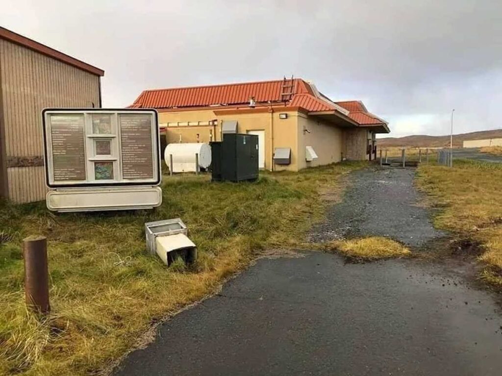 An abandoned McDonald's drive-thru with overgrown grass and a weathered menu sign. The building has a red-tiled roof and peeling paint, and the parking area is cracked and empty.