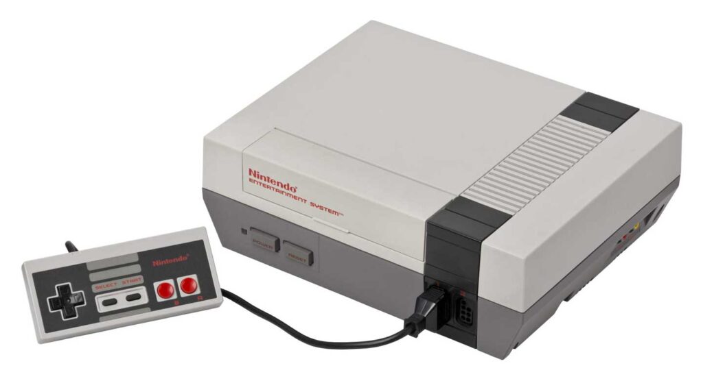 A classic Nintendo Entertainment System console, one of the best-selling video game consoles, features a grey rectangular design connected to a matching controller. The controller has a black D-pad and red buttons, perfectly placed beside the console on a pristine white background.