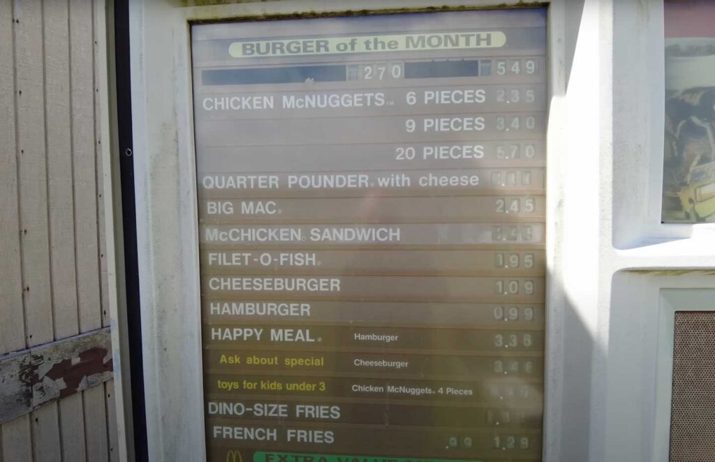 A fast food drive-thru menu displays various items and prices, including the Burger of the Month, Chicken McNuggets, Big Mac, McChicken Sandwich, Filet-O-Fish, Cheeseburgers, and Happy Meals. Prices are listed beside each item.