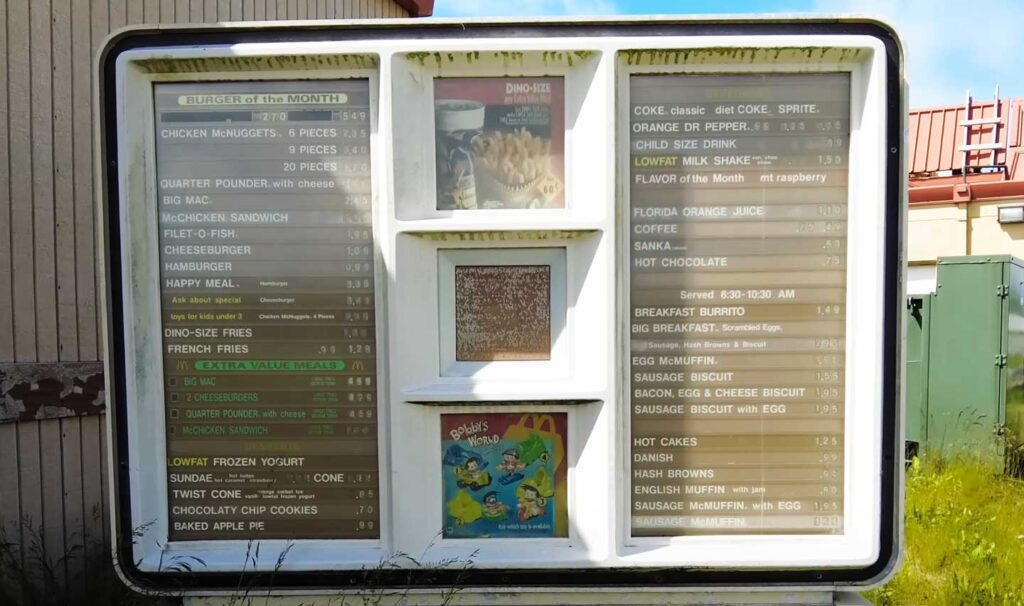 A vintage McDonald's drive-thru in Adak Alaska menu board displaying various items such as burgers, chicken nuggets, breakfast meals, and drinks. The menu includes a Burger of the Month section. The board is outdoors and slightly weathered.